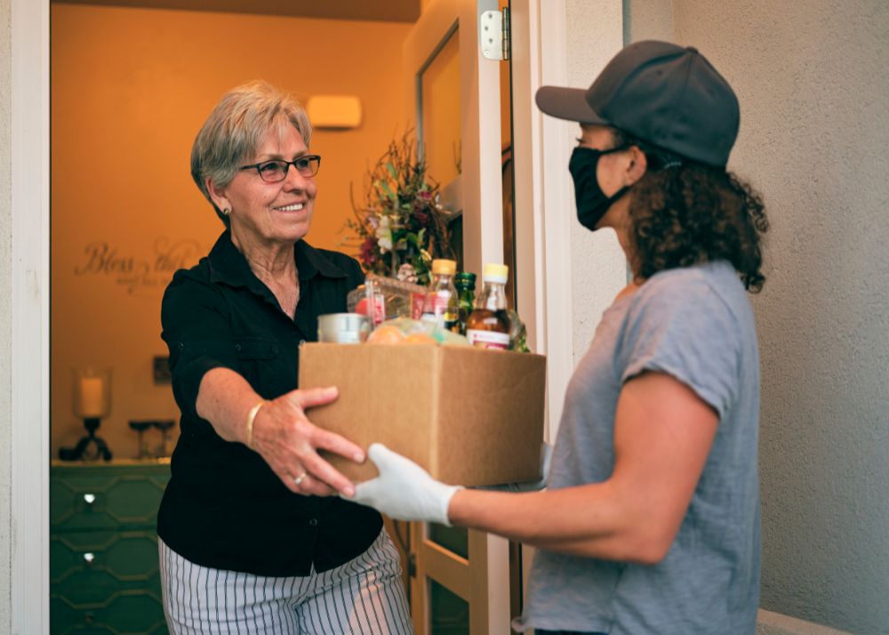 Displayed is a delivery person with a filtered mask delivering a box of fresh groceries to an older woman at her frontdoor.