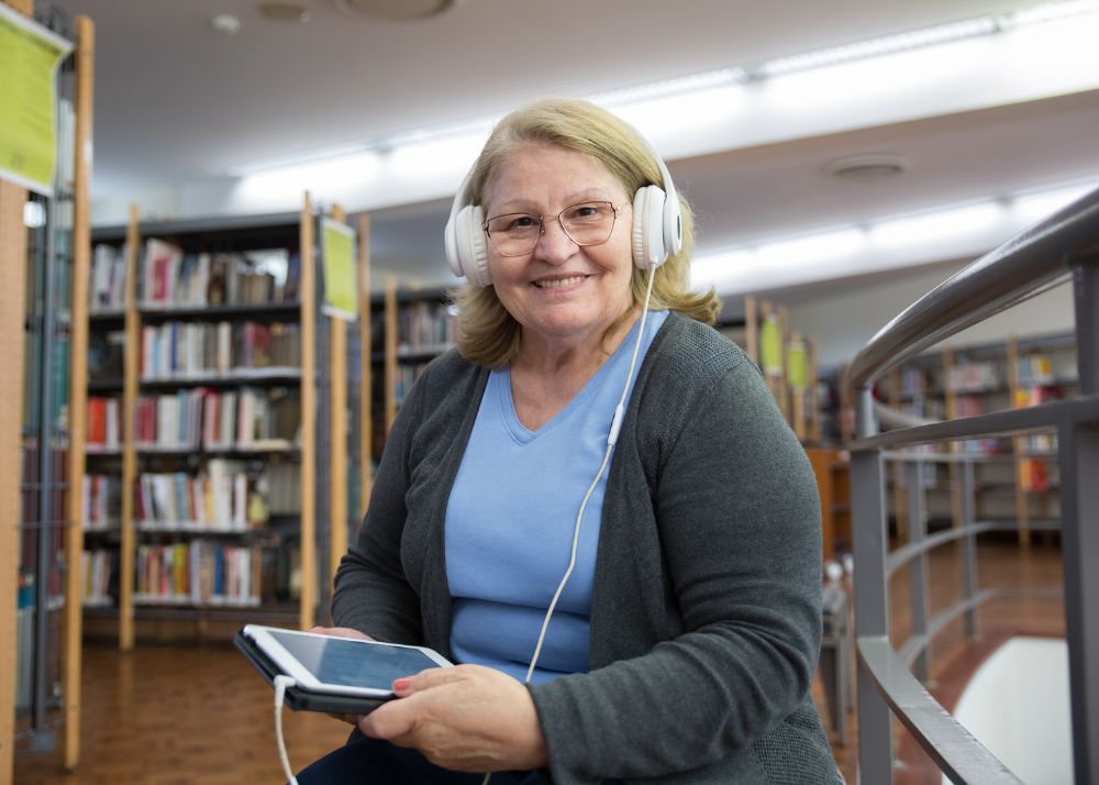 Displayed is an older woman in a library, holding a tablet and wearing headphones that are attached to the tablet.